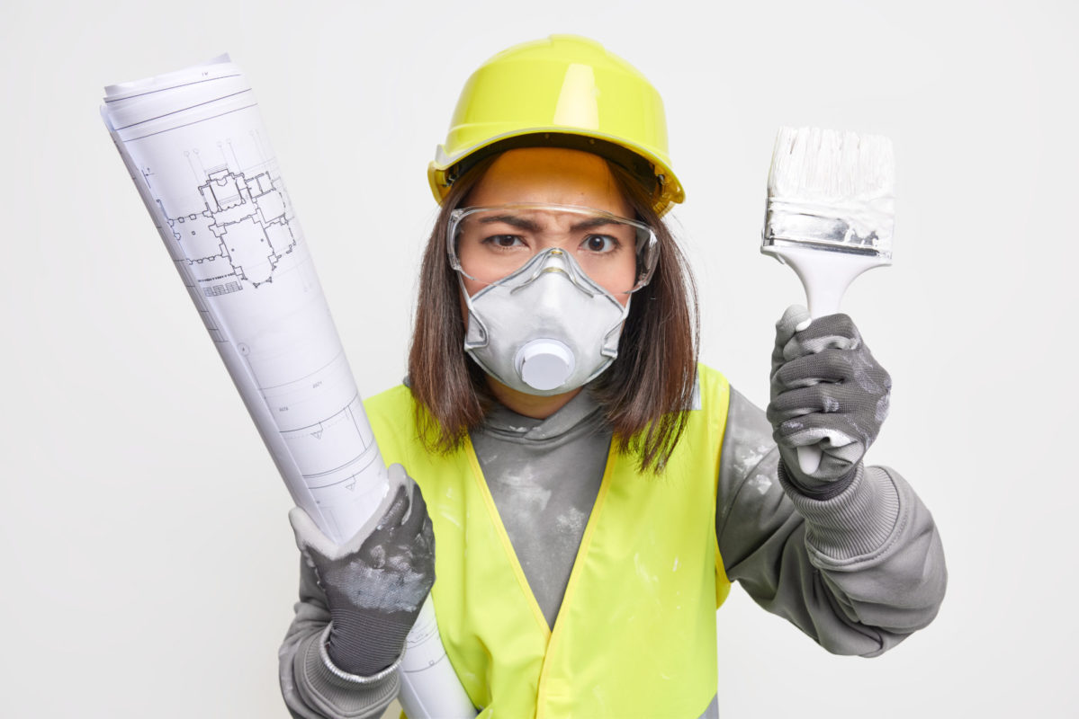 photo of serious strict female builder works on construction site prepares architectural plannings holds building blueprint and paint brush wears protective uniform